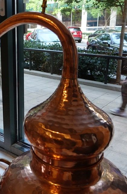 A hammered copper pot still with a swan neck, lit by daylight from the tasting-room window